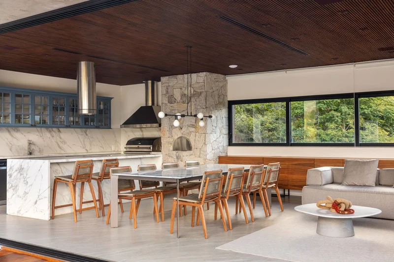 Área gourmet moderna e integrada com a piscina, com churrasqueira de inox, bancada de ilha e móveis de madeira.